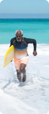 Photo of a senior man running through the shallow water on a tropical beach with a surfboard under his arm.