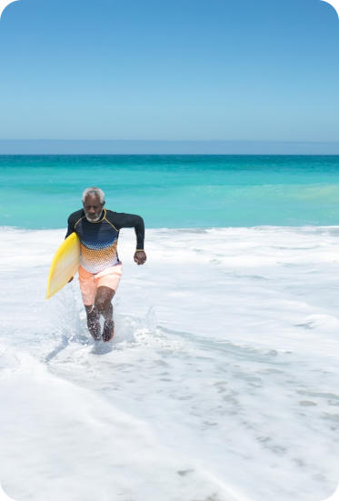 Photo of a senior man running through the shallow water on a tropical beach with a surfboard under his arm.