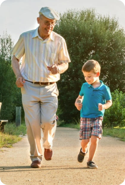 A senior man jogging happily down a gravel road on a sunny day with a young child running next to him.