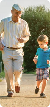 A senior man jogging happily down a gravel road on a sunny day with a young child running next to him.